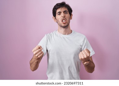 Young hispanic man standing over pink background angry and mad raising fists frustrated and furious while shouting with anger. rage and aggressive concept.  - Shutterstock ID 2320697311
