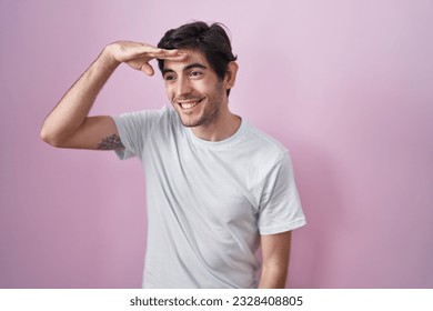 Young hispanic man standing over pink background very happy and smiling looking far away with hand over head. searching concept.  - Shutterstock ID 2328408805
