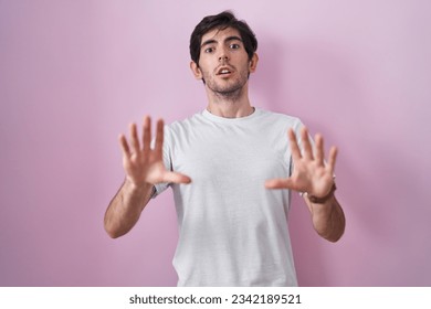 Young hispanic man standing over pink background afraid and terrified with fear expression stop gesture with hands, shouting in shock. panic concept. 