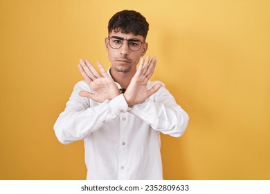 Young hispanic man standing over yellow background rejection expression crossing arms and palms doing negative sign, angry face  - Shutterstock ID 2352809633