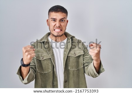 Young hispanic man standing over isolated background angry and mad raising fists frustrated and furious while shouting with anger. rage and aggressive concept. 