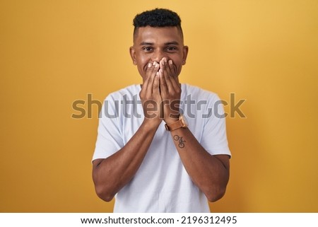 Young hispanic man standing over yellow background laughing and embarrassed giggle covering mouth with hands, gossip and scandal concept 