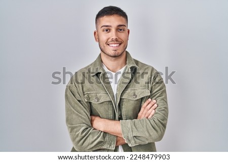 Young hispanic man standing over isolated background happy face smiling with crossed arms looking at the camera. positive person. 