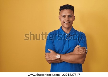 Young hispanic man standing over yellow background happy face smiling with crossed arms looking at the camera. positive person. 