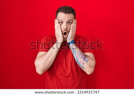 Young hispanic man standing over red background afraid and shocked, surprise and amazed expression with hands on face 