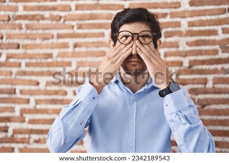 Young hispanic man standing over brick wall background rubbing eyes for fatigue and headache, sleepy and tired expression. vision problem 
