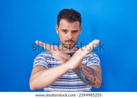 Young hispanic man standing over blue background rejection expression crossing arms doing negative sign, angry face 