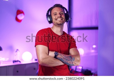 Young hispanic man streamer smiling confident standing with arms crossed gesture at gaming room