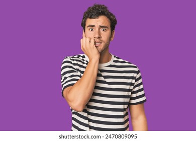 Young Hispanic man in studio biting fingernails, nervous and very anxious.