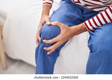 Young Hispanic Man Suffering For Knee Injury Sitting On Bed At Bedroom