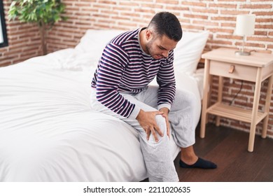 Young Hispanic Man Suffering For Knee Injury Sitting On Bed At Bedroom