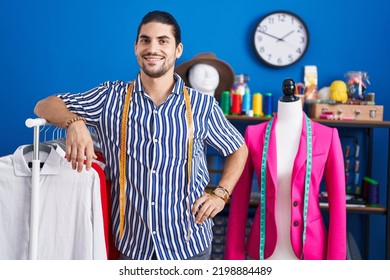 Young Hispanic Man Tailor Smiling Confident Leaning On Clothes Rack At Sewing Studio