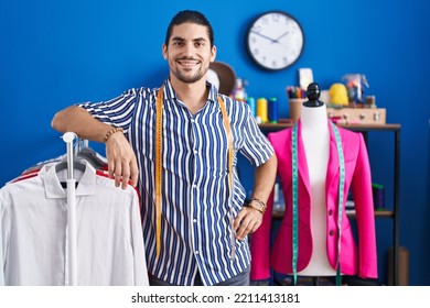 Young Hispanic Man Tailor Smiling Confident Leaning On Clothes Rack At Sewing Studio