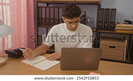Young hispanic man taking notes while working on a laptop in an office with bookshelves in the background.