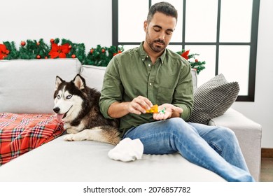 Young Hispanic Man Taking Pills Sitting On Sofa With Dog By Christmas Decor At Home