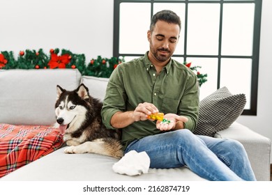 Young Hispanic Man Taking Pills Sitting On Sofa With Dog By Christmas Decor At Home