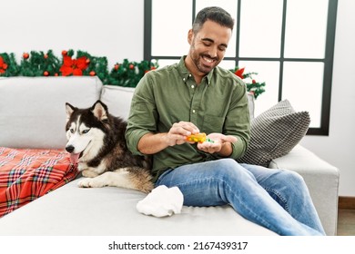 Young Hispanic Man Taking Pills Sitting On Sofa With Dog By Christmas Decor At Home