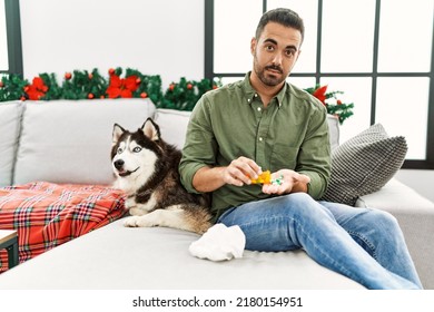 Young Hispanic Man Taking Pills Sitting On Sofa With Dog By Christmas Decor At Home