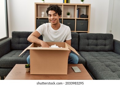 Young Hispanic Man Unboxing Cardboard Box Sitting On The Sofa At Home.