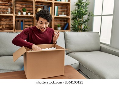 Young Hispanic Man Unboxing Cardboard Box Sitting On The Sofa At Home.