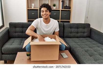 Young Hispanic Man Unboxing Cardboard Box Sitting On The Sofa At Home.