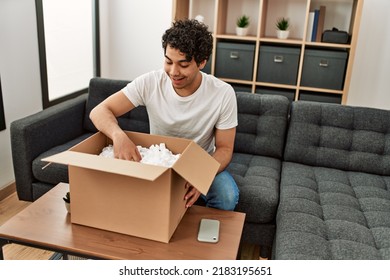 Young Hispanic Man Unboxing Cardboard Box Sitting On The Sofa At Home.