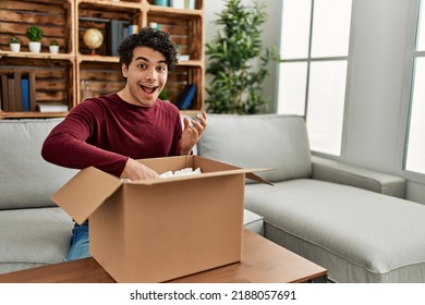 Young Hispanic Man Unboxing Cardboard Box Sitting On The Sofa At Home.