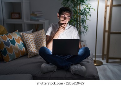 Young Hispanic Man Using Laptop At Home At Night With Hand On Chin Thinking About Question, Pensive Expression. Smiling With Thoughtful Face. Doubt Concept. 