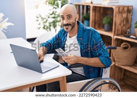 Young hispanic man using laptop holdig dollars sitting on wheelchair at home