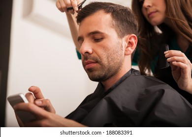 Young Hispanic Man Using A Smart Phone While Getting A Haircut At A Hair Salon