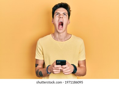 Young Hispanic Man Using Smartphone Typing Message Angry And Mad Screaming Frustrated And Furious, Shouting With Anger Looking Up. 