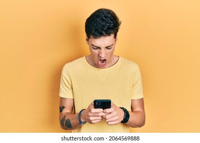 Young Hispanic Man Using Smartphone Typing Message Angry And Mad Screaming Frustrated And Furious, Shouting With Anger. Rage And Aggressive Concept. 