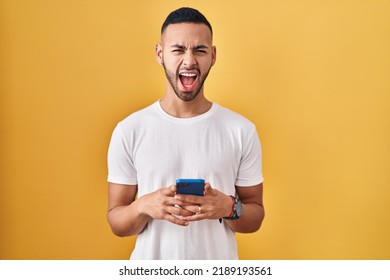 Young Hispanic Man Using Smartphone Typing Message Angry And Mad Screaming Frustrated And Furious, Shouting With Anger. Rage And Aggressive Concept. 