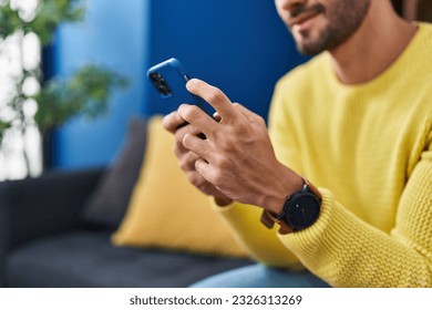 Young hispanic man using smartphone sitting on sofa at home