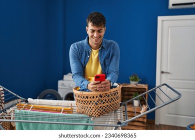 Young hispanic man using smartphone hanging clothes on clothesline at laundry room