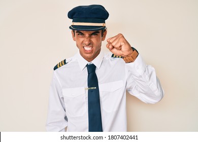 Young Hispanic Man Wearing Airplane Pilot Uniform Angry And Mad Raising Fist Frustrated And Furious While Shouting With Anger. Rage And Aggressive Concept. 