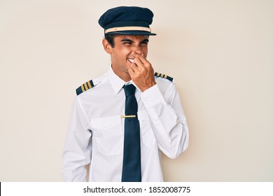 Young Hispanic Man Wearing Airplane Pilot Uniform Smelling Something Stinky And Disgusting, Intolerable Smell, Holding Breath With Fingers On Nose. Bad Smell 