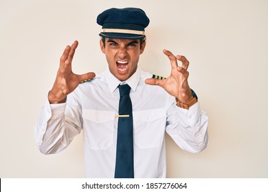 Young Hispanic Man Wearing Airplane Pilot Uniform Shouting Frustrated With Rage, Hands Trying To Strangle, Yelling Mad 