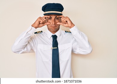 Young Hispanic Man Wearing Airplane Pilot Uniform Trying To Open Eyes With Fingers, Sleepy And Tired For Morning Fatigue 