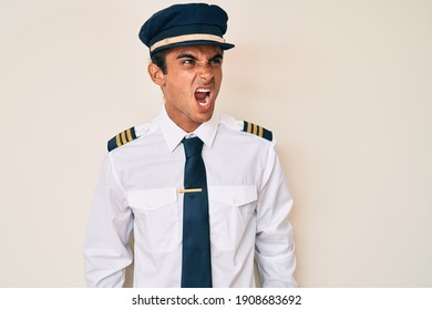 Young Hispanic Man Wearing Airplane Pilot Uniform Angry And Mad Screaming Frustrated And Furious, Shouting With Anger. Rage And Aggressive Concept. 