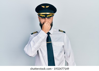 Young Hispanic Man Wearing Airplane Pilot Uniform Smelling Something Stinky And Disgusting, Intolerable Smell, Holding Breath With Fingers On Nose. Bad Smell 