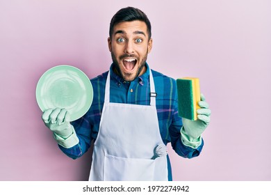 Young Hispanic Man Wearing Apron Holding Scourer Washing Dishes Celebrating Crazy And Amazed For Success With Open Eyes Screaming Excited. 