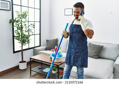 Young Hispanic Man Wearing Apron And Gloves Talking On The Smartphone At Home