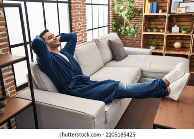 Young Hispanic Man Wearing Bathrobe Relaxed With Hands On Head At Home