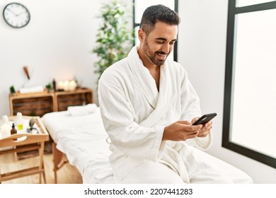 Young Hispanic Man Wearing Bathrobe Using Smartphone At Beauty Center