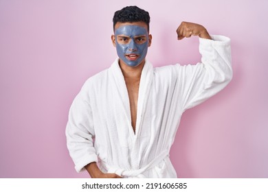 Young Hispanic Man Wearing Beauty Face Mask And Bath Robe Strong Person Showing Arm Muscle, Confident And Proud Of Power 