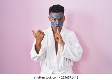 Young hispanic man wearing beauty face mask and bath robe asking to be quiet with finger on lips pointing with hand to the side. silence and secret concept. 