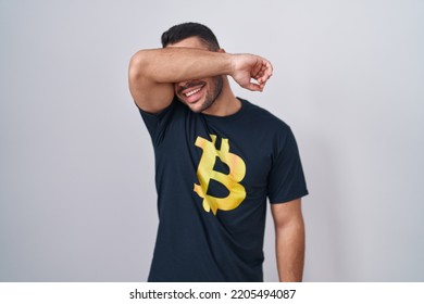 Young Hispanic Man Wearing Bitcoin T Shirt Smiling Cheerful Playing Peek A Boo With Hands Showing Face. Surprised And Exited 