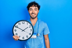 Young Hispanic Man Wearing Blue Male Nurse Uniform Holding Clock Looking Positive And Happy Standing And Smiling With A Confident Smile Showing Teeth 