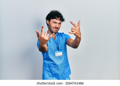 Young Hispanic Man Wearing Blue Male Nurse Uniform Shouting With Crazy Expression Doing Rock Symbol With Hands Up. Music Star. Heavy Concept. 